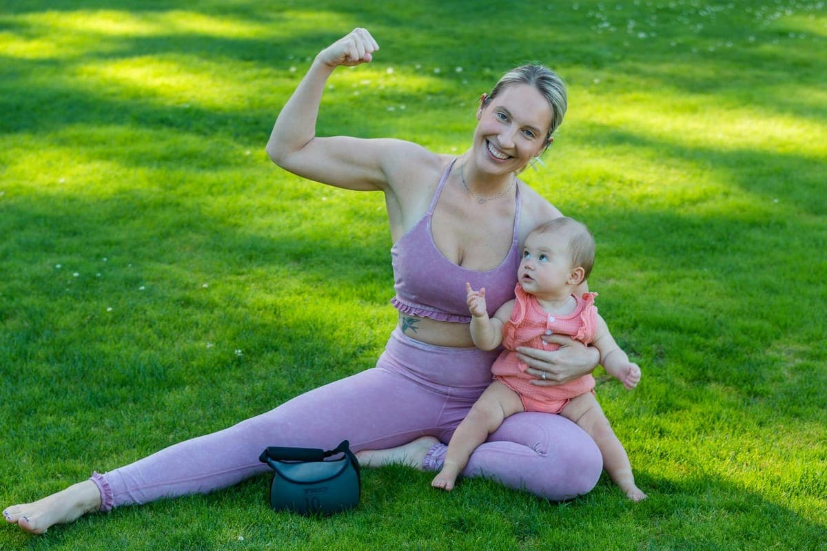 A tired but smiling new mum eating a healthy snack one-handed while holding her newborn baby